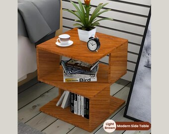 May include: A light brown modern side table with a three-tiered design. The top shelf holds a small white teacup and saucer, a black alarm clock, and a potted plant. Books are visible on the lower shelves. The table is approximately 24 inches tall.