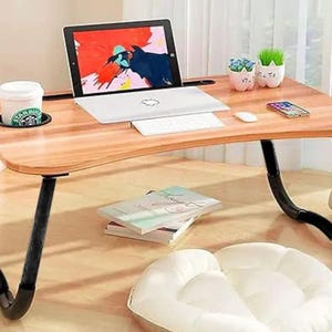 May include: A wooden laptop desk with black legs, a built-in cup holder, and a slot for a tablet. A laptop, keyboard, mouse, and phone are on the desk. Decorative plants and a coffee cup are also visible. The desk is designed for use on a bed or floor.
