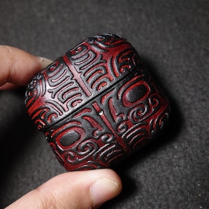 May include: A square-shaped protective case for wireless earbuds featuring a deep red and black color scheme. The surface is decorated with an intricate, embossed pattern of swirling lines and geometric shapes that cover the entire exterior.