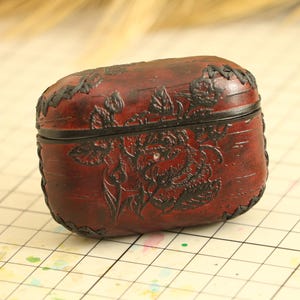Könnte beinhalten: Eine rechteckige, weinrote Lederbox mit Klappdeckel. Die Box hat ein detailliertes geprägtes Blumendesign und schwarze Nähte an den Kanten. Auf der Vorderseite der Box ist ein kleiner, silberfarbener Niet zu sehen.