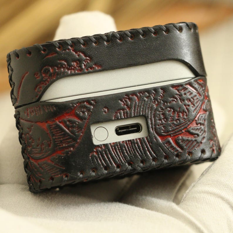 May include: Black leather AirPods case with red floral embossed design. The case has a stitched border and a cutout for the charging port. The case is designed to protect the AirPods.