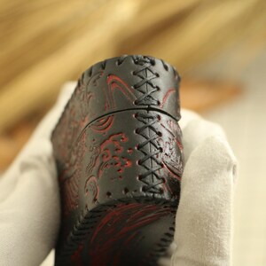 May include: Black leather case with red embossed wave pattern and cross-stitched seams. The case has a cylindrical shape and is held by a gloved hand. The leather has a textured appearance.