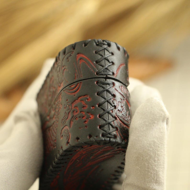 May include: Black leather case with red embossed wave pattern and cross-stitched seams. The case has a cylindrical shape and is held by a gloved hand. The leather has a textured appearance.