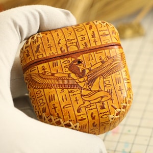 以下が含まれることがあります： 黄色の革製のケースで、茶色のステッチの縁取りとエジプトの女神のデザインが施されています。ケースにはヒエログリフが印刷されています。