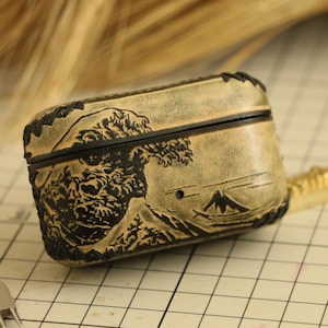 以下が含まれることがあります： 黒い蓋とトリムが付いたベージュ色の革製ケース。ケースには、波と山のシーンの詳細なレリーフが施されています。デザインは日本の芸術を彷彿とさせます。ケースは長方形で、小型電子機器用と思われます。