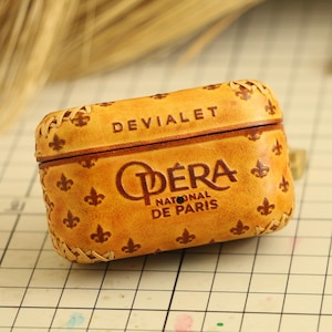 以下が含まれることがあります： 上面に「DEVIALET」の文字が入った、タン色の革製ケース。ケースの前面には「OPERA NATIONAL DE PARIS」の文字と、フルール・ド・リスの模様があります。ケースにはステッチが施されています。