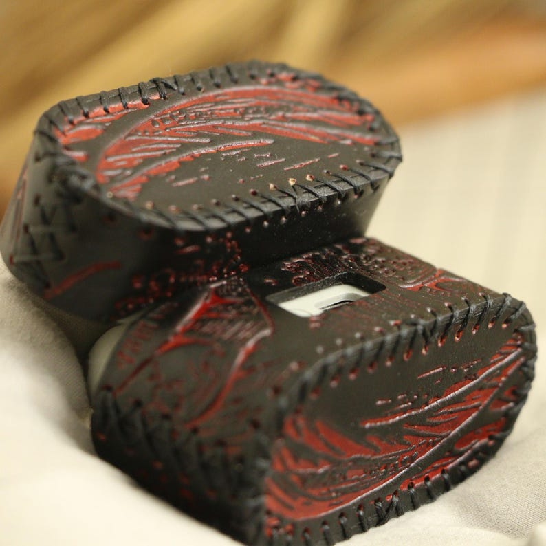 May include: A black leather case with red decorative detailing and stitching. The case is rectangular with rounded edges and a textured surface. The red detailing appears to be carved or embossed into the leather, creating a detailed design.