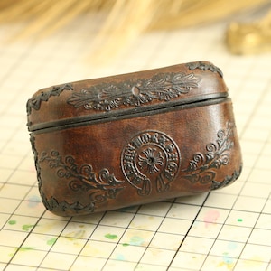 以下が含まれることがあります： 黒の複雑な花柄と十字架のデザインが施された茶色の革ケース。ケースはステッチされた縁とフラップが付いています。