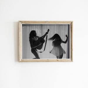 以下が含まれることがあります： 2人のシルエットが描かれた白黒の額入りプリント。一人はギターを弾き、もう一人は踊っています。プリントは明るい色の木製フレームに入っており、ミニマリストな美学を作り出しています。