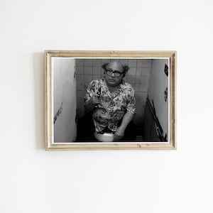 May include: Black and white framed print featuring a man in a floral shirt and glasses holding a gun while sitting on a toilet. The background shows a tiled wall and a narrow space.