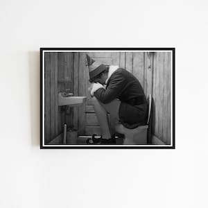 May include: Black and white framed print of a person dressed as an elf, sitting on a toilet in a rustic bathroom. The elf is wearing a green hat, a dark jacket, and black shoes. A small sink and toilet brush are visible.