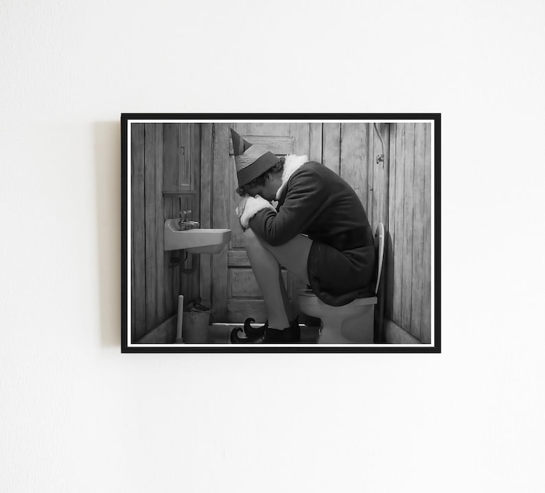 May include: Black and white framed print of a person dressed as an elf, sitting on a toilet in a rustic bathroom. The elf is wearing a green hat, a dark jacket, and black shoes. A small sink and toilet brush are visible.