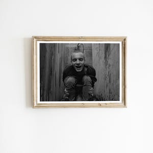 Op de afbeelding: Zwart-wit ingelijste foto van een hurkende persoon. De persoon draagt een donker shirt en jeans, met een verraste uitdrukking. De achtergrond is een houten constructie.