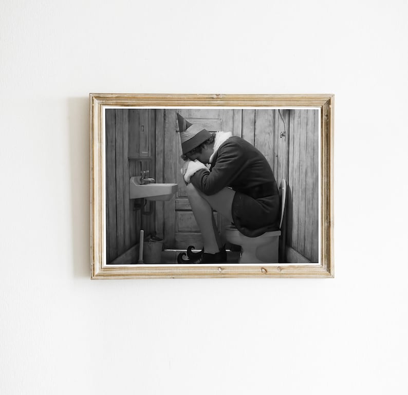 May include: Black and white framed print of a person dressed as an elf, sitting on a toilet with their head in their hands. The elf is wearing a green and red hat, a dark jacket, and black shoes.