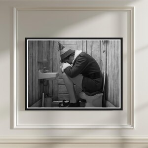 May include: Black and white framed print of a person dressed as an elf, sitting on a toilet in a rustic bathroom. The elf is wearing a hat, coat, and elf shoes. A small sink and bucket are visible.