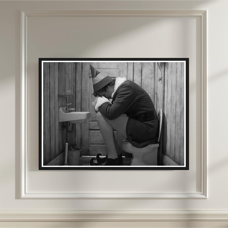 May include: Black and white framed print of a person dressed as an elf, sitting on a toilet in a rustic bathroom. The elf is wearing a hat, coat, and elf shoes. A small sink and bucket are visible.