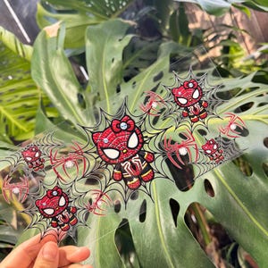 May include: A clear, rectangular sticker sheet featuring a cartoon character mashup. The design combines elements of Spider-Man and Hello Kitty, with red, black, and white colors. The sheet includes multiple images of the character on spiderwebs, with rhinestone accents.