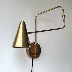Peut inclure: Une applique murale en laiton avec un abat-jour conique et un bras réglable. La lampe a une base circulaire et une articulation rectangulaire. Un cordon noir part de la base. La lampe est en laiton.