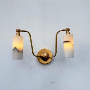 Puede incluir: Lámpara de pared con dos pantallas cilíndricas. Las pantallas son blancas con vetas grises y negras, y emiten un brillo cálido. La lámpara tiene un acabado de latón y un diseño curvo.