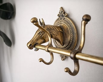 Könnte beinhalten: Ein wandmontierter Kleiderhaken in Messingfarbe mit einem Pferdekopf-Design. Der Haken hat eine horizontale Stange und drei Haken. Der Pferdekopf ist im Profil dargestellt, mit detaillierten Merkmalen und einer dekorativen runden Rückseite.