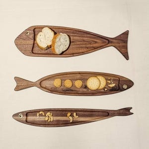 May include: Three wooden serving trays shaped like fish, each with a recessed area. The top tray holds bread slices, the middle tray has crackers, and the bottom tray contains cashews. The trays are made of dark wood.