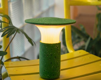 Puede incluir: Una lámpara de exterior verde moteada con una parte superior plana y redonda y una base cilíndrica. La lámpara emite una luz cálida y amarilla. La lámpara está sobre un banco amarillo.
