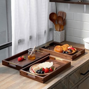 May include: Three rectangular wooden serving trays with gold handles are arranged on a wooden surface. The trays hold food items like strawberries, honey, and pastries. A jar of honey with a wooden stick is visible.