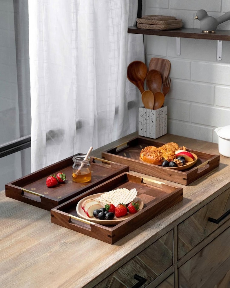 May include: Three rectangular wooden serving trays with gold handles are arranged on a wooden surface. The trays hold food items like strawberries, honey, and pastries. A jar of honey with a wooden stick is visible.