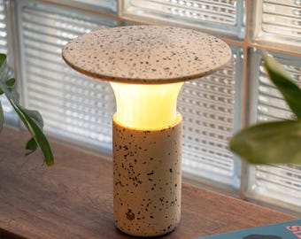 May include: A modern, mushroom-shaped table lamp with a speckled, off-white shade and base. The lamp emits a warm, yellow glow. The cylindrical base and the top are made of a speckled material. The lamp is sitting on a wooden surface.