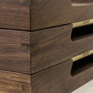 May include: Close-up of three stacked, dark brown wooden trays. Each tray features a rectangular shape with a recessed handle and a small, gold-colored label. The wood grain is visible, and the joinery is expertly crafted.