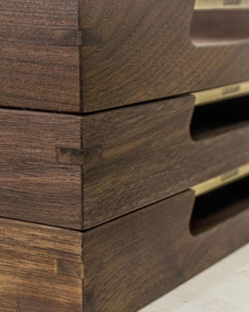 May include: Close-up of three stacked, dark brown wooden trays. Each tray features a rectangular shape with a recessed handle and a small, gold-colored label. The wood grain is visible, and the joinery is expertly crafted.