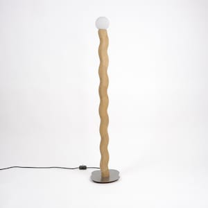 Puede incluir: Una lámpara de pie moderna con un poste de madera ondulado de color claro y una bombilla redonda blanca esmerilada en la parte superior. La base es de forma irregular y color plateado. Un cable de alimentación negro sale de la base.