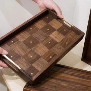 May include: A rectangular wooden serving tray with a checkered pattern and brass accents. The tray is dark brown with lighter wood squares and small brass squares. The handles are brass. Another tray is visible in the background.