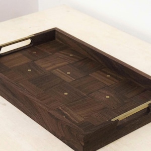 May include: A rectangular wooden serving tray with a dark brown, parquet-style surface. The tray has brass-colored handles and small brass accents. The tray is designed for serving food or drinks.