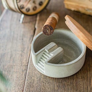 May include: A gray ceramic ashtray with a built-in cigar rest, a lit cigar, and a wooden cigar holder on a wooden surface. The ashtray has a stepped design. The cigar is brown with a white band.