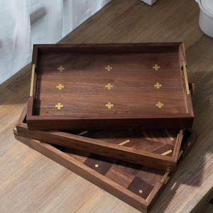 May include: Three wooden serving trays stacked on a wooden surface. The top tray is rectangular with brass handles and gold cross inlays. The middle tray has a geometric design with gold accents. The bottom tray has a square pattern with gold accents.