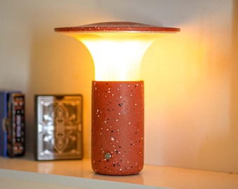 May include: A terracotta-colored table lamp with a speckled pattern. The lamp has a cylindrical base and a flared top, emitting a warm, inviting glow. The lamp is on a white shelf, with a deck of cards and a book in the background.