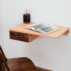 Könnte beinhalten: Ein an der Wand befestigter Holzschreibtisch mit einer Schublade und einem Buch. Der Schreibtisch besteht aus hellem Holz mit einer dunklen Holzschublade und einem Messinggriff. Ein schwarz-goldener Stifthalter und ein Holzstuhl sind ebenfalls zu sehen.