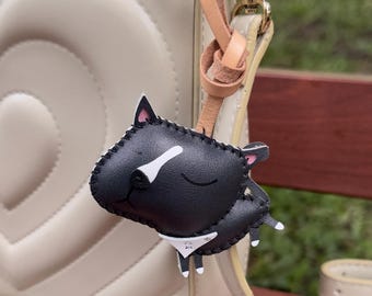 May include: A black leather cat keychain with white accents, including a white nose and a small white bib. The cat has pink ears and is attached to a tan leather strap and gold-colored hardware. The keychain is hanging from a cream-colored purse.