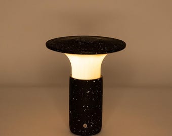 May include: A modern black and white speckled table lamp with a mushroom-shaped top. The lamp has a cylindrical base and a warm, glowing light emanating from the center. The lamp is approximately 12 inches tall.