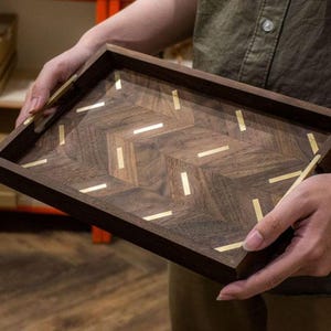 May include: A rectangular wooden tray with a dark brown finish and a herringbone pattern. The tray is inlaid with brass accents. The tray is being held by a person. The tray is a decorative item.