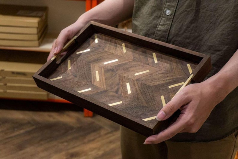 May include: A rectangular wooden tray with a dark brown finish and a herringbone pattern. The tray is inlaid with brass accents. The tray is being held by a person. The tray is a decorative item.