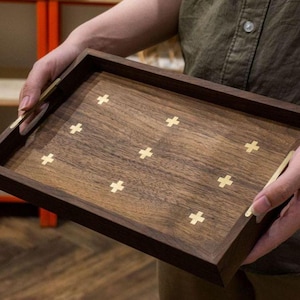 May include: A rectangular wooden tray with a dark brown frame and a lighter brown interior. The tray features inlaid gold-colored cross designs. The tray is being held by a person.