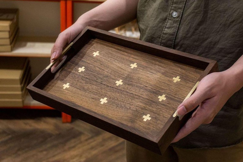 May include: A rectangular wooden tray with a dark brown frame and a lighter brown interior. The tray features inlaid gold-colored cross designs. The tray is being held by a person.