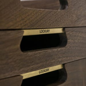 May include: Close-up of three stacked wooden serving trays. Each tray is a dark brown color with a rectangular shape and a cutout handle. A gold-colored metal band with the word "LOOXURY" is affixed to each tray.