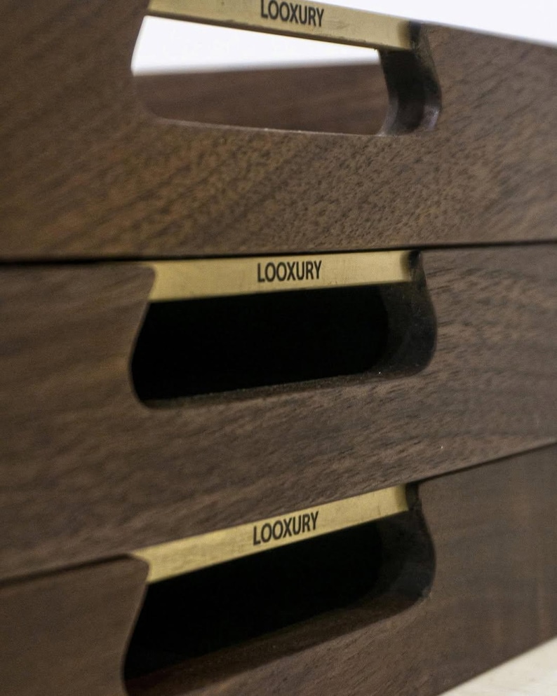 May include: Close-up of three stacked wooden serving trays. Each tray is a dark brown color with a rectangular shape and a cutout handle. A gold-colored metal band with the word "LOOXURY" is affixed to each tray.