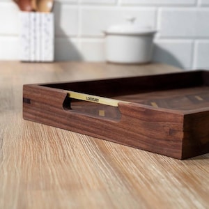 May include: A rectangular wooden serving tray with a dark brown finish. The tray has a gold-colored handle with the word "LOOKXURY" printed on it. The tray is on a light-colored wooden surface.