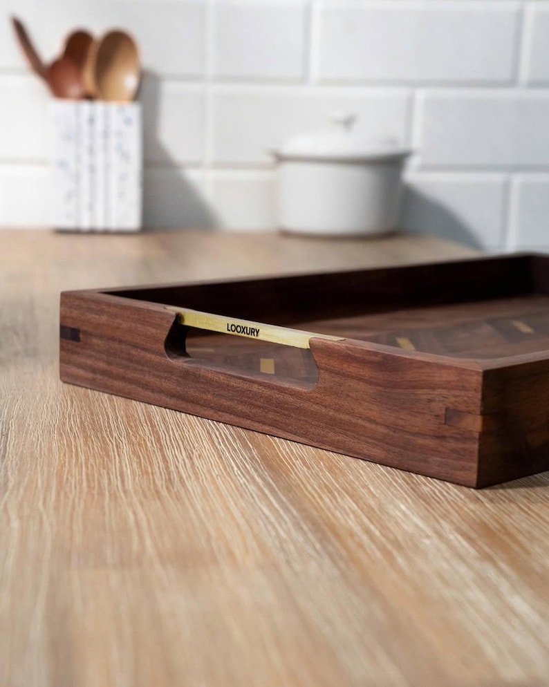 May include: A rectangular wooden serving tray with a dark brown finish. The tray has a gold-colored handle with the word "LOOKXURY" printed on it. The tray is on a light-colored wooden surface.