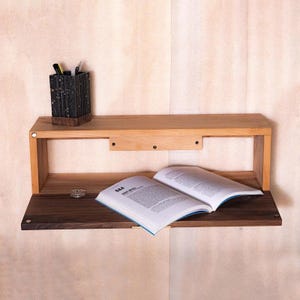 Puede incluir: Estantería flotante de madera de dos niveles con acabado natural. La balda superior contiene un portalápices y la balda inferior un libro abierto. La estantería está montada en una pared de color claro.