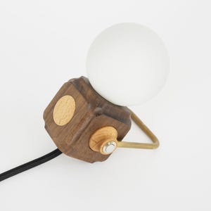 Op de afbeelding: Een moderne tafellamp met een witte globe lamp op een donkere houten kubus met een licht houten accent. De lamp heeft een messing arm en een zwart snoer. Het ontwerp van de lamp is minimalistisch en eigentijds.
